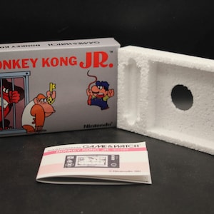 May include: A vintage Donkey Kong Jr. Game & Watch box set, including the game's box, a white foam insert, and an instruction manual. The box displays the game's title and cartoon graphics of Donkey Kong Jr. and other characters.