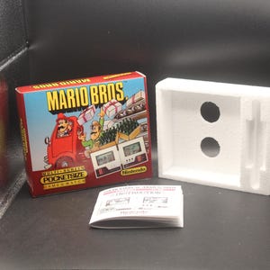 May include: A boxed Nintendo Mario Bros. PocketSize Game & Watch. The box features a red truck with Mario and Luigi, and the game's title. A white foam insert and instruction manual are also present.