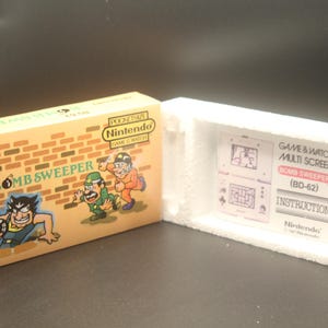 Könnte beinhalten: Eine gelbe und braune Schachtel mit einem Ziegelmuster und dem Text "BOMB SWEEPER" auf der Vorderseite. Die Schachtel enthält ein Game & Watch Multi Screen "BOMB SWEEPER" (BD-62) Handheld-Spiel. Die Schachtel ist geöffnet und das Spiel ist im Inneren sichtbar.