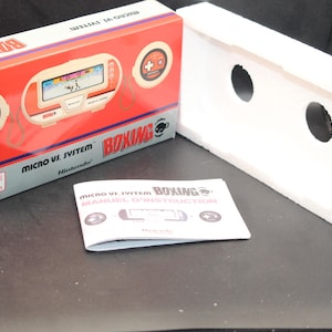 Puede incluir: Una consola de videojuegos portátil roja y blanca con temática de boxeo. La consola tiene una pantalla que muestra un combate de boxeo. La caja del juego es roja y blanca con las palabras "Micro VS System" y "Boxing" impresas en ella. El juego está hecho por Nintendo.