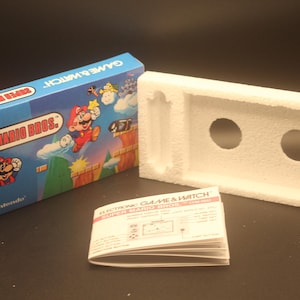 Op de afbeelding: Een blauwe en witte doos met de tekst "GAME & WATCH SUPER MARIO BROS." en een afbeelding van Mario die springt. De doos ligt naast een witte styropor verpakking en een gevouwen instructieboekje met de tekst "ELECTRONIC GAME & WATCH SUPER MARIO BROS. INSTRUCTION MANUAL".