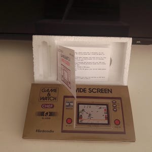 Może przedstawiać: Zabytkowa gra przenośna Nintendo Game & Watch Chef Wide Screen w oryginalnym opakowaniu. Brązowe pudełko wyświetla nazwę i funkcje gry. Ekran gry pokazuje grę o tematyce kulinarnej z timerem i wynikiem.