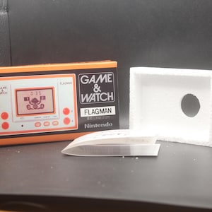 Op de afbeelding: Een oranje en zwarte Game & Watch Flagman handheld console van Nintendo. De doos is open en toont de console, een witte schuimrubberen inzet en een doorzichtige plastic folie. Het scherm toont een karakter.