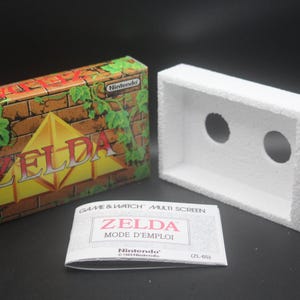 Puede incluir: Una consola de juegos portátil Game & Watch Zelda vintage en su caja original. La caja es marrón con un patrón de pared de ladrillo y enredaderas verdes. El cartucho del juego es blanco con dos agujeros para la pantalla.