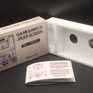 Puede incluir: Una caja blanca con el texto "GAME & WATCH MULTI SCREEN OIL PANIC" y el logotipo de Nintendo. La caja contiene una consola de juegos portátil Game & Watch. La consola es una versión de pantalla múltiple del juego "Oil Panic".