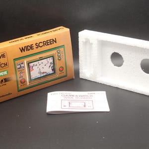 May include: A vintage Game & Watch Wide Screen handheld game console box, featuring the "Popeye" game. The tan box has green and red details. A white polystyrene insert and instruction manual are also visible.