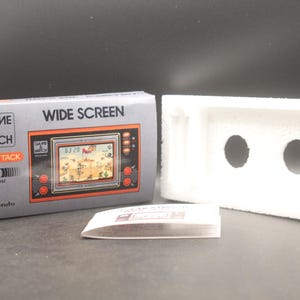 Könnte beinhalten: Eine graue Schachtel für ein Nintendo Game & Watch Wide Screen Fire Attack Spiel. Die Schachtel trägt den Spieletitel und ein Bild des Spielbildschirms. Ein weißer Schaumstoffeinsatz und ein kleines Merkblatt sind ebenfalls vorhanden.