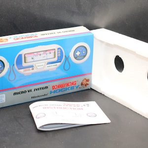 Puede incluir: Una consola de juegos portátil Nintendo Game & Watch blanca y azul con un juego de Donkey Kong Hockey. La consola está en su caja original con un inserto de espuma blanca. La caja tiene un fondo azul con las palabras "Donkey Kong Hockey" y "Micro VS. System" impresas en ella.