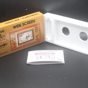Peut inclure: Jeu portable Nintendo Game & Watch Wide Screen MC-25 Mickey Mouse vintage dans sa boîte d'origine. La boîte est beige avec des accents rouges et noirs, et l'écran du jeu montre Mickey Mouse. Comprend un insert en mousse blanc et un manuel d'instructions.