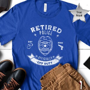 May include: A royal blue t-shirt with a white graphic design that says "Retired Police" with a badge, handcuffs, a gun, and the text "Officially Off Duty".