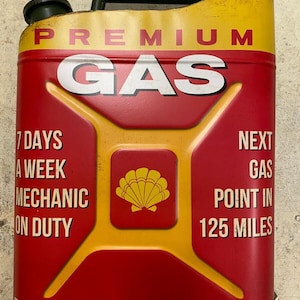 May include: A red and yellow metal gas can with the words "PREMIUM GAS" and "MOTOR OIL" printed on it. The can has a black handle and a black cap. It also has a yellow seashell logo and the text "7 DAYS A WEEK MECHANIC ON DUTY" and "NEXT GAS POINT IN 125 MILES".