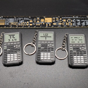 Op de afbeelding: Drie rekenmachine sleutelhangers met zilveren sleutelringen en kettingen. De rekenmachines tonen "π 3.14", "∫" en een reeks streepjes. Een zwarte liniaal met gouden tekst staat boven de sleutelhangers.