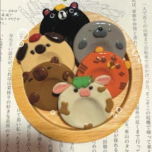 以下が含まれることがあります： 様々な動物を模した、形と色が異なるお守り石のコレクション。クマ、アザラシ、トラ、ウシ、イヌの形に作られています。木製のプレートに配置され、上部には「Worry Stone」の文字があります。