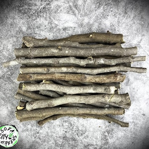 May include: A pile of small, natural wooden sticks in varying shades of brown and gray. The sticks are arranged in a haphazard stack, with some extending outwards. The background is a textured, gray surface. The sticks appear to be dry and have a rough texture.