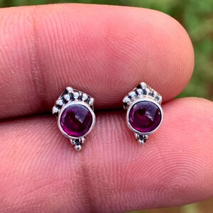 Könnte beinhalten: Ein Paar kleiner Ohrstecker aus Silber. Jeder Ohrring hat einen runden, tiefroten Edelstein, der in eine dekorative Silberfassung eingesetzt ist. Die Ohrringe haben ein detailliertes, verziertes Design um den Edelstein herum.
