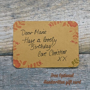 Free Optional handwritten gift card. Brown recycled craft card with a border of red flowers and leaves Art Deco style. Handwritten message from customer to friend: Dear Marie, Have a lovely birthday! love Christine XX. Background of blue grey wood.