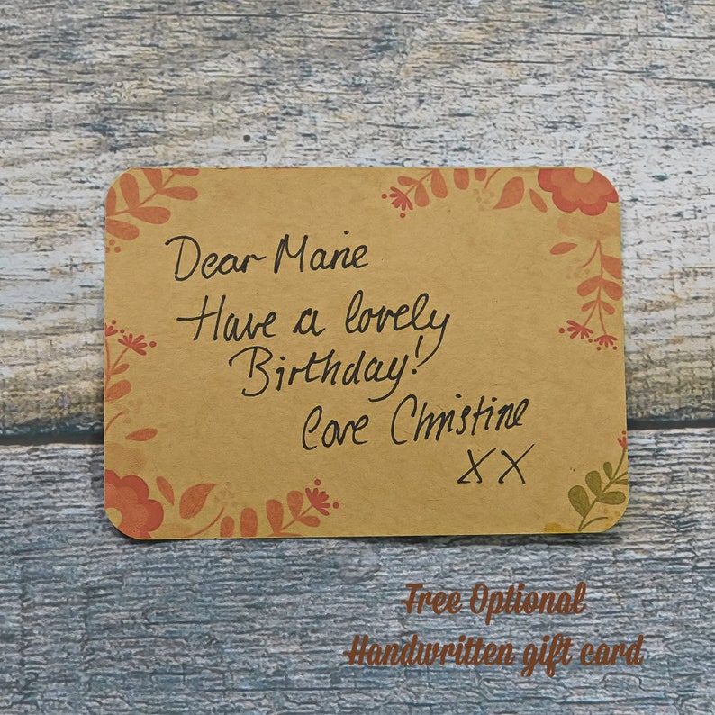 Free Optional handwritten gift card. Brown recycled craft card with a border of red flowers and leaves Art Deco style. Handwritten message from customer to friend: Dear Marie, Have a lovely birthday! love Christine XX. Background of blue grey wood.