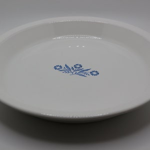 May include: White pie plate with a blue floral design. The design features three blue flowers with green stems and leaves.
