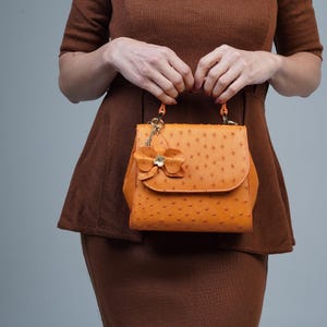 Può includere: Borsa a mano in pelle arancione con un ciondolo a forma di fiore. La borsa ha una superficie strutturata e una chiusura con patta. La borsa è tenuta da una persona che indossa un vestito marrone. La borsa ha un manico superiore e una catena dorata.