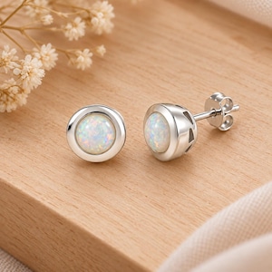 May include: A pair of stud earrings featuring round, iridescent white opal cabochons set in polished silver-toned metal. Each earring has a smooth, circular bezel setting and a standard post back with a butterfly clutch mechanism for secure wear.