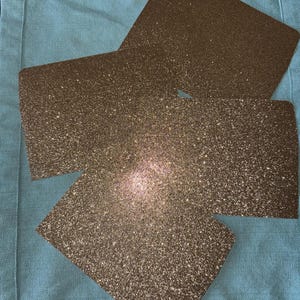 May include: Three gold glitter sheets on a light blue fabric background.