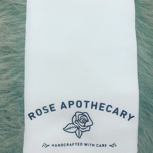 May include: White cotton tea towel with black text that reads "ROSE APOTHECARY" and a black line drawing of a rose. The text "HANDCRAFTED WITH CARE" is also printed in black.