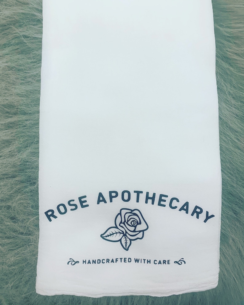 May include: White cotton tea towel with black text that reads "ROSE APOTHECARY" and a black line drawing of a rose. The text "HANDCRAFTED WITH CARE" is also printed in black.