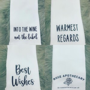 May include: Four white tea towels with black text. The first towel says "INTO THE WINE not the label". The second towel says "WARMEST REGARDS". The third towel says "Best Wishes". The fourth towel says "ROSE APOTHECARY" with a small rose graphic and the text "HANDCRAFTED WITH CARE".