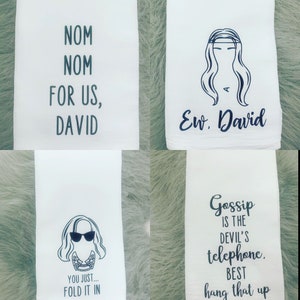 May include: Four white tea towels with black text and illustrations. The first towel says "Nom Nom For Us, David". The second towel has a black illustration of a woman with long hair and the text "Ew, David". The third towel has a black illustration of a woman with sunglasses and the text "You Just... Fold It In". The fourth towel says "Gossip Is The Devil's Telephone. Best Hang That Up".