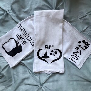 May include: Three white kitchen towels with black text and graphics. The first towel says "Unadulterated Loafing" with a slice of bread graphic. The second towel says "BFF" with a heart graphic containing a witch's hat and wand. The third towel says "Popu-lar" with a wand graphic.