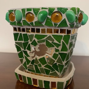 Peut inclure: Un pot de fleurs en mosaïque vert avec une silhouette de grenouille et une bordure dorée. Le pot a une soucoupe et est décoré de carreaux de verre verts et dorés.