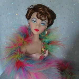 May include: A doll with brown hair and red lipstick wearing a pink dress and a colorful feather boa.