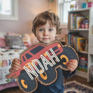 Op de afbeelding: Een kind houdt een rode en zwarte monstertruck-uitknipsel vast met de naam "NOAH" erop geschreven in witte letters. Het uitknipsel is gemaakt van hout en heeft een verweerde afwerking.