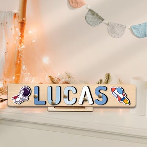 May include: Wooden puzzle with the name "LUCAS" in white and blue letters. The puzzle features an astronaut and a rocket ship graphic. The puzzle is displayed on a wooden stand.