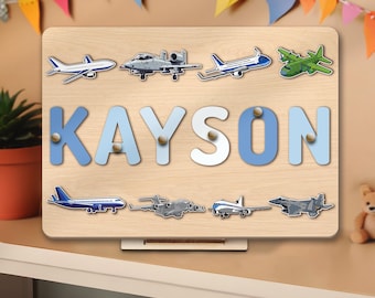 May include: A wooden personalised puzzle with the name Kayson in blue and white letters. The puzzle features various aeroplane images, including a fighter jet, a cargo plane, and a passenger plane.