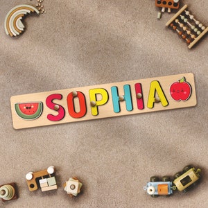May include: A wooden name puzzle featuring the name "Sophia" spelled out in colourful letters. The puzzle also includes a watermelon and an apple.