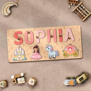 May include: A wooden puzzle featuring the name "SOPHIA" spelled out in pink letters. The puzzle includes a princess, a unicorn, a castle, and a carriage. The puzzle is set against a light brown background.