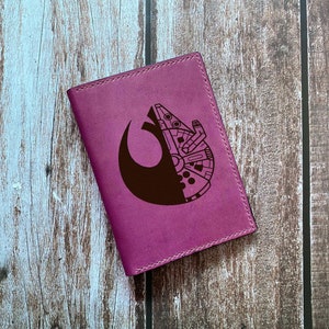 Op de afbeelding: Een paarse leren paspoorthouder met een zwart-wit ontwerp van de Millennium Falcon en het Rebel Alliance-symbool.