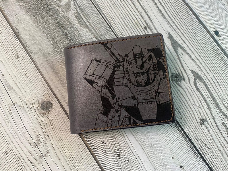 May include: A brown leather wallet with a black and white graphic of a robot. The robot has a large head with a visor and a large body with arms and legs.