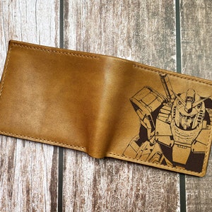 May include: A brown leather wallet with a laser-engraved image of a robot. The robot is in a standing position with a large head and a weapon in its right hand.
