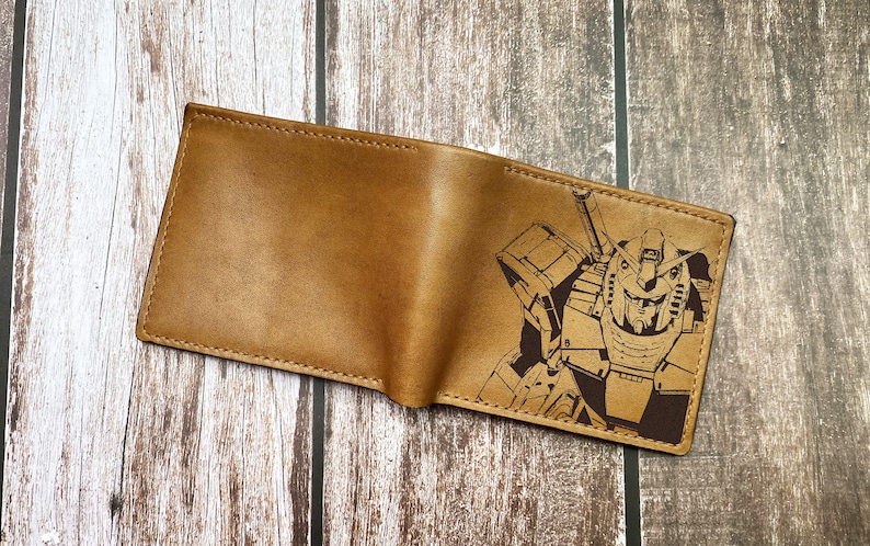 May include: A brown leather wallet with a laser-engraved image of a robot. The robot is in a standing position with a large head and a weapon in its right hand.