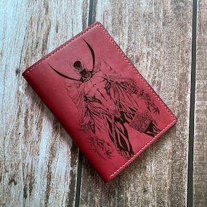 May include: A red leather wallet featuring a detailed black illustration of a comic book character. The wallet has a stitched border and a rectangular shape. The artwork depicts a figure in a cape and mask.