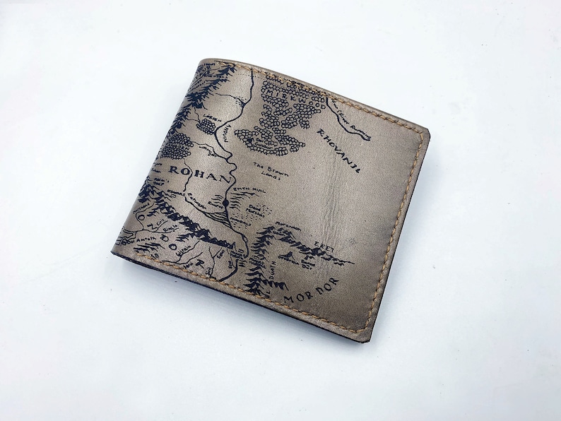 May include: A brown leather wallet with a map of Middle-earth from The Lord of the Rings printed on the front. The map includes the names of locations like Rohan, Mordor, and Rhovanion.