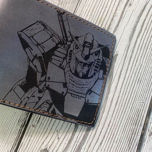 May include: A gray leather wallet with a black and white graphic of a robot printed on the front. The wallet has brown stitching around the edges.