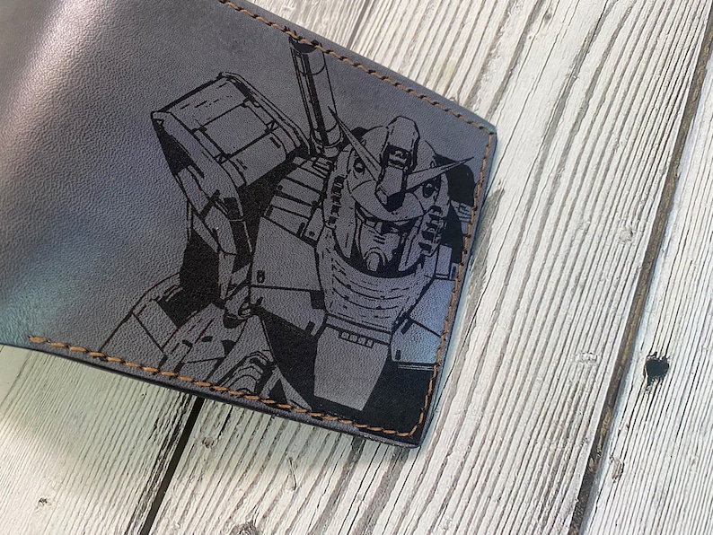 May include: A gray leather wallet with a black and white graphic of a robot printed on the front. The wallet has brown stitching around the edges.