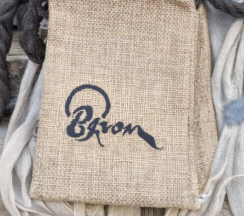 May include: A beige burlap bag with the word "Bqron" in dark blue script. The bag is surrounded by gray and beige fabric and rope.