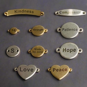 Może przedstawiać: Zestaw metalowych zawieszek z różnymi wygrawerowanymi słowami i frazami, w tym "Kindness", "Compassion", "Strength", "Patience", "Hope", "Love", "Peace" i "Made by Jane".