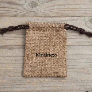May include: A small, rectangular burlap bag with a drawstring closure. The bag is tan with the word "Kindness" printed in black. The drawstring is a dark brown color. The bag is sitting on a light brown wooden surface.