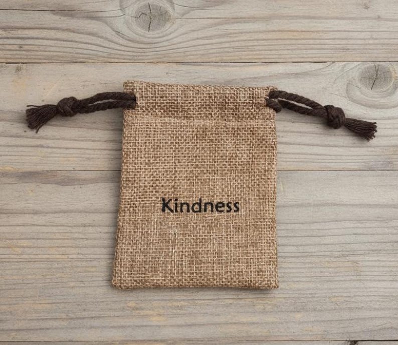 May include: A small, rectangular burlap bag with a drawstring closure. The bag is tan with the word "Kindness" printed in black. The drawstring is a dark brown color. The bag is sitting on a light brown wooden surface.