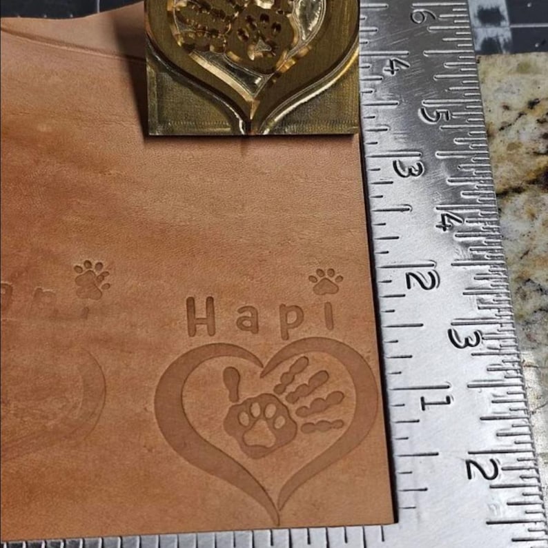 May include: A brown leather piece with the word "Hapi" and a paw print inside a heart. There are two paw prints on the left side of the image.